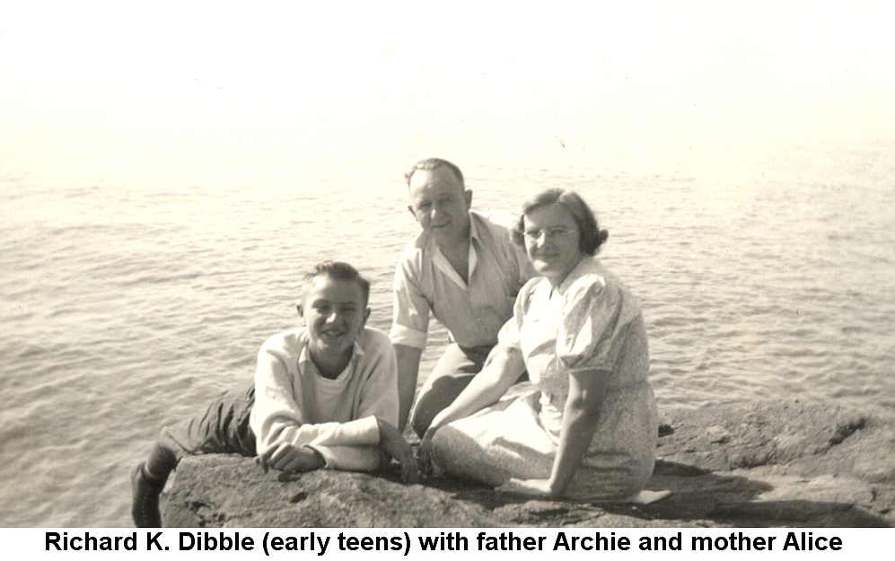 Black and white photo of teen-aged Richard K. Dibble wearing a light-colored sweater, white t-shirt, dark pants and shoes, lying semi-prone on a large rock outcropping, propped up on his elbows and smiling. To his right his father Archie kneels on the rock, leaning on his right hand; Alice in a white dress and glasses, with short curled hair, sits on the rock next to them. The calm water and bright sky stretch away from them into the distance.